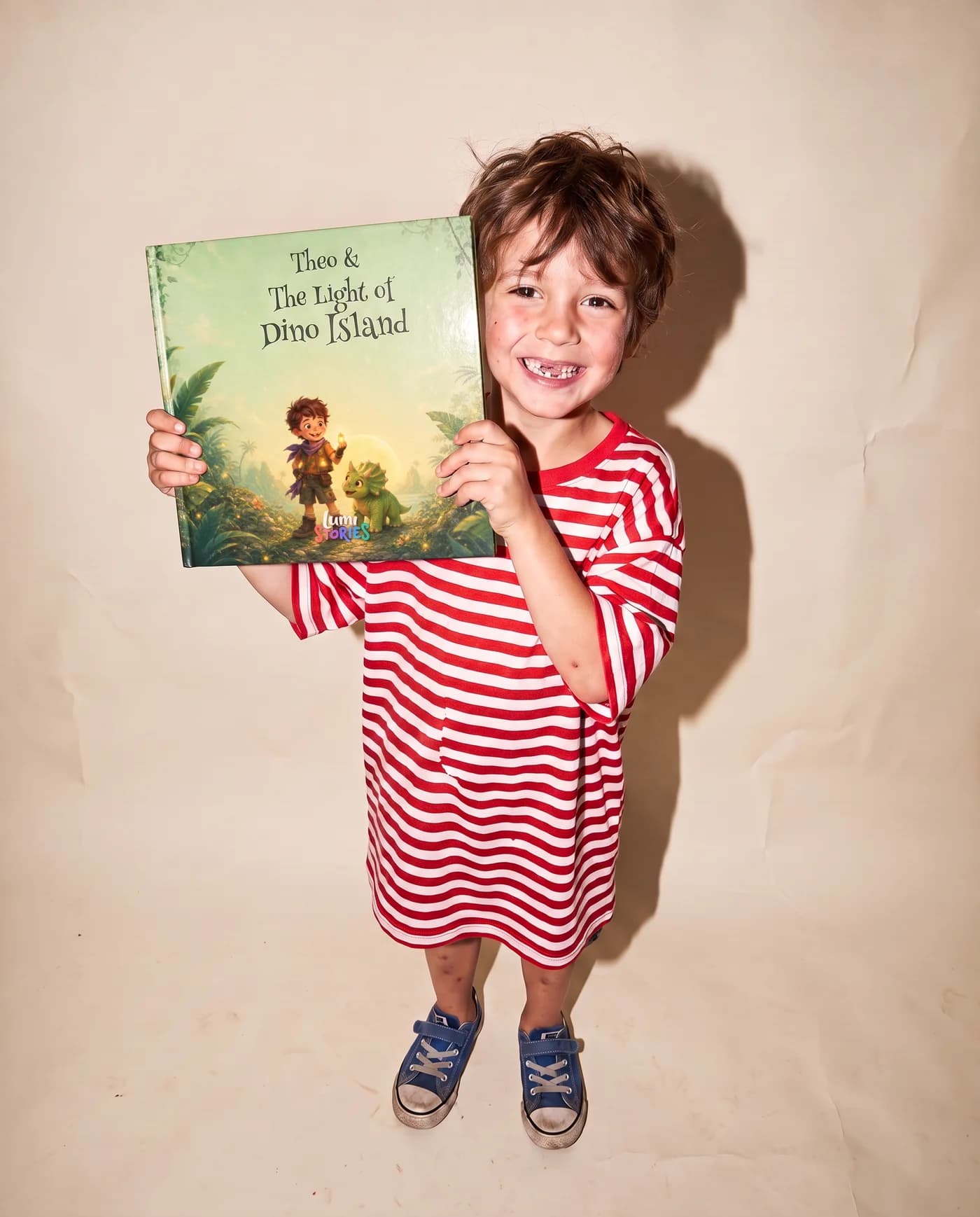 A boy on a studio backdrop holds up his own copy, Theo and the Light of Dino Island, with his illustrated hero on the cover