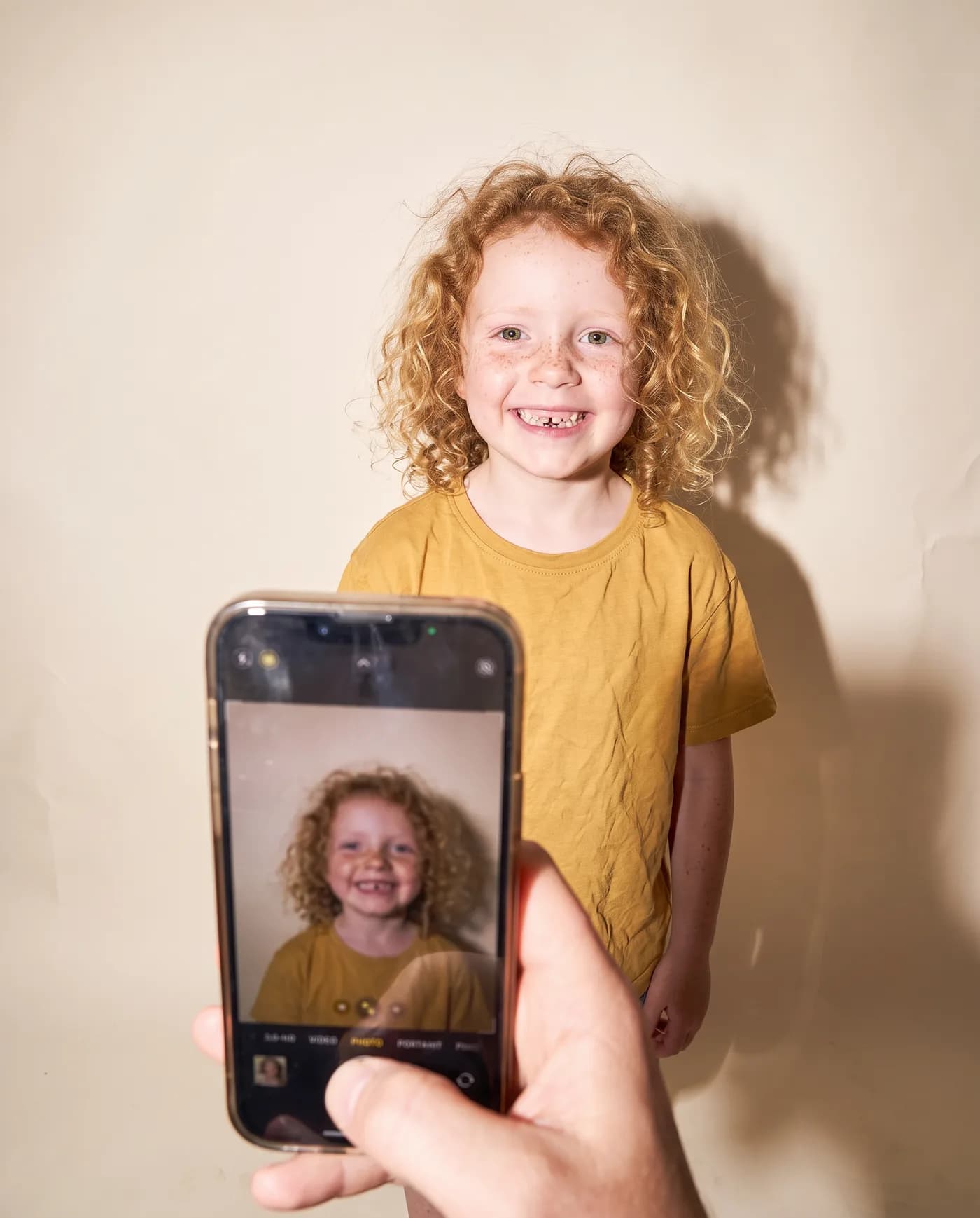 A parent’s phone photographs a grinning girl with curly golden hair on a studio backdrop