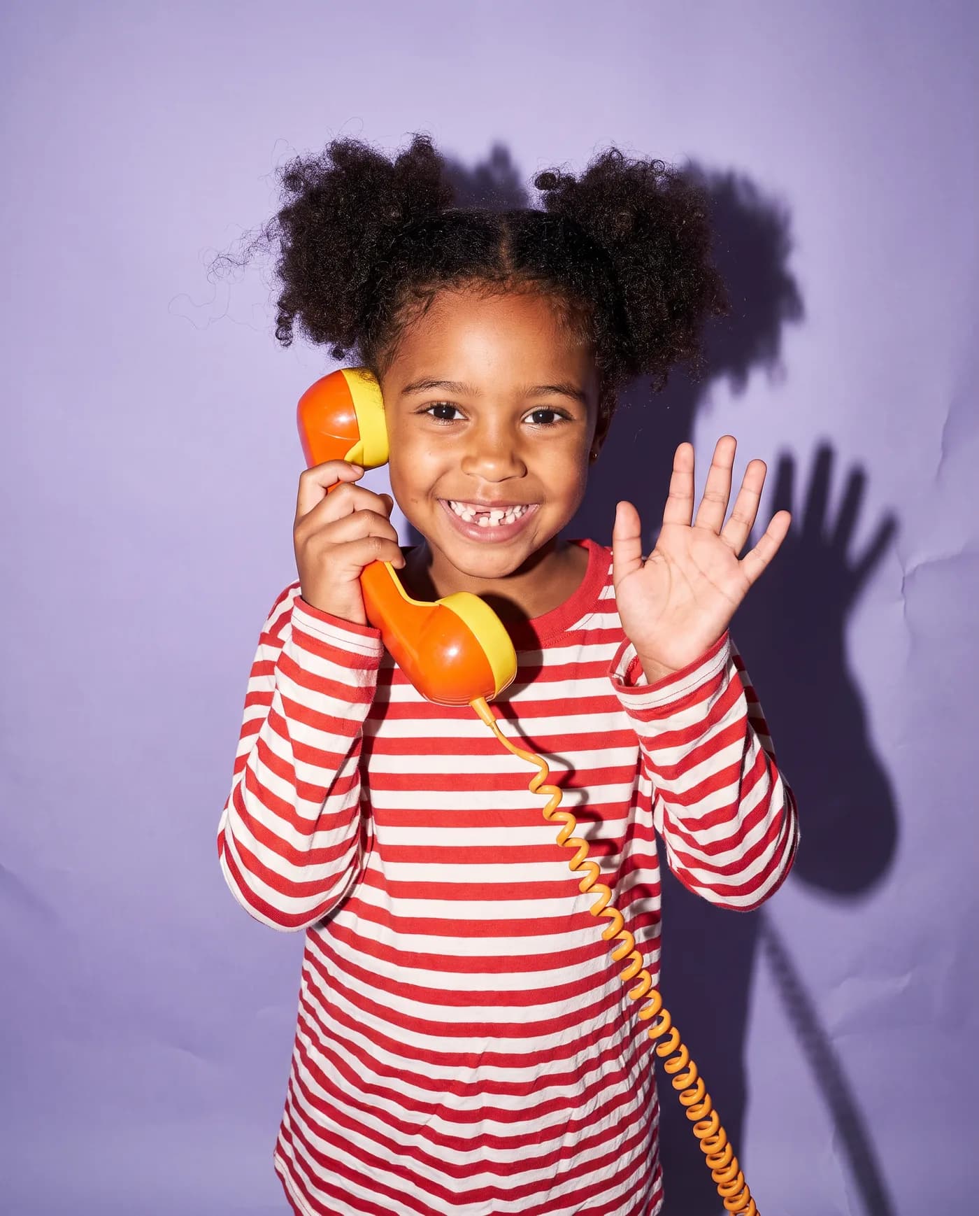 A girl on a lavender backdrop waves while talking on a toy phone