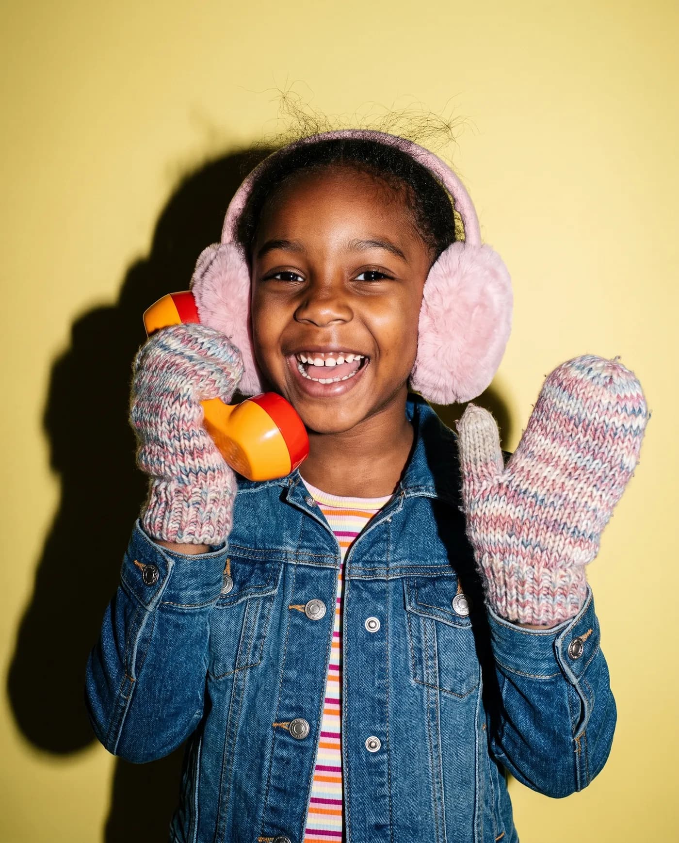 A girl in earmuffs waves while talking on a toy phone