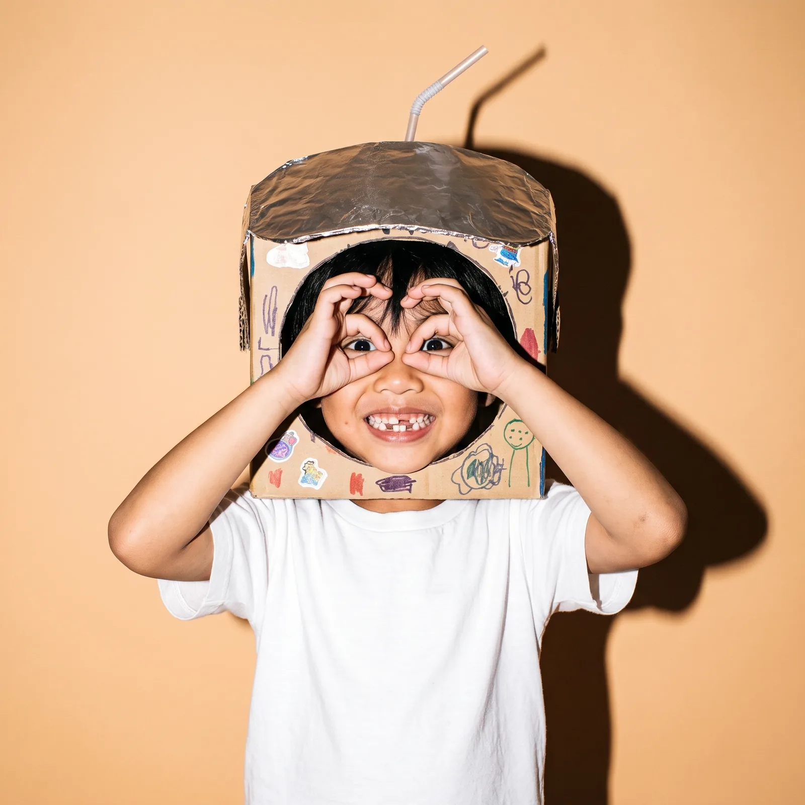 A boy in a cardboard astronaut helmet makes goggles with his hands on a peach backdrop